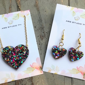 Op de afbeelding: Een set hartvormige sieraden met een ketting en oorbellen. De hartvormige hanger en oorbellen zijn gemaakt van hars en gevuld met iriserende glitter. De ketting heeft een gouden ketting en de oorbellen hebben gouden haakjes. De sieraden worden gepresenteerd op witte kaartjes met de tekst "ARS STUDIO 74".