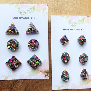 Op de afbeelding: Twee sets kleurrijke glitterhars oorbellen in verschillende vormen, waaronder cirkels, driehoeken, vierkanten, diamanten en druppelvormen. De oorbellen worden gepresenteerd op witte kaarten met de tekst "ARS STUDIO 74".