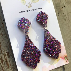 May include: A pair of dangle earrings with a purple, pink, and blue glitter finish. The earrings are shaped like a seashell and have a teardrop-shaped dangle. The earrings are on a white card with the text "ARS STUDIO 74".