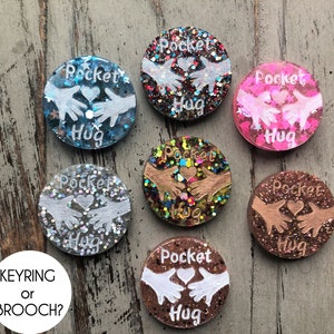 May include: Seven round resin keychains or brooches with glitter embedded in the resin. Each keychain features a white silhouette of two hands holding a heart with the text "Pocket Hug" in white lettering.