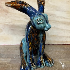 May include: A ceramic hare sculpture featuring a glossy glaze in shades of deep blue, light blue, and amber brown. The hare is depicted in a seated position with long, upright ears, a detailed face, and visible paw structures on its front legs.