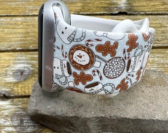 May include: A light blue Apple Watch strap with a festive holiday pattern. The strap features snowmen, gingerbread men, candy canes, and coffee cups in brown and white. The watch strap is shown on a grey stone.