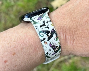 Puede incluir: Correa de reloj blanca con un diseño de Halloween. La correa presenta murciélagos, fantasmas, estrellas y sombreros de bruja en negro, morado, verde y blanco. La caja del reloj es negra.