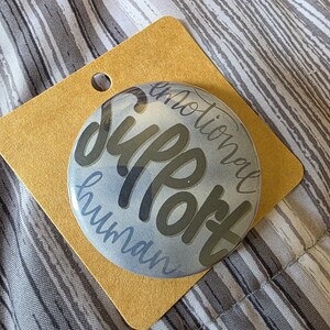 May include: A circular pinback button featuring a light blue and grey background with the text "emotional SUPPORT human" printed in a dark grey, stylized script font. The button is attached to a square, tan-colored cardboard display backing.