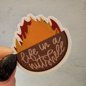 May include: A white sticker with a brown and orange illustration of a flame inside a nutshell. The text "life in a nutshell" is written in brown cursive.