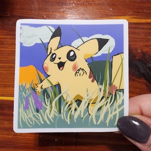 May include: Square sticker featuring Pikachu, a yellow creature with black-tipped ears and red cheeks, in a grassy field. The background shows a blue sky with clouds and a mountain.