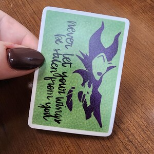 May include: A green sticker with a silhouette of Maleficent from the Disney movie "Sleeping Beauty". The sticker has the text "Never let your wings be stolen from you".