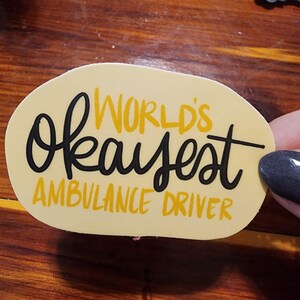 May include: A yellow oval sticker with black text that reads "World's okayest ambulance driver".
