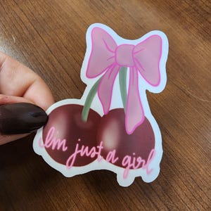 May include: A white sticker with a pink bow and two cherries. The text "I'm just a girl" is written in pink on the sticker.