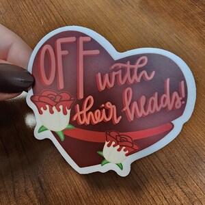 May include: A red heart-shaped sticker with the text "OFF with their heads!" in white and red lettering. The sticker has two red roses with white centers and green leaves.