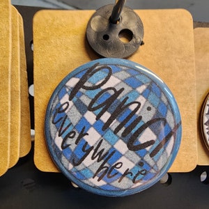 May include: A blue and white plaid button with the text "Panic everywhere" written in black.