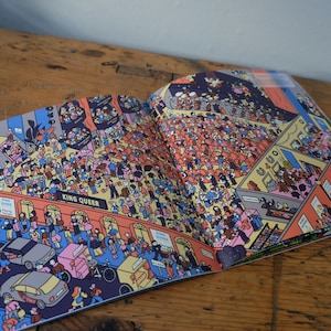 Może przedstawiać: Otwarta książka z kolorową ilustracją zatłoczonego pokoju. Ilustracja jest pełna ludzi, samochod&oacute;w i budynk&oacute;w. Na książce widoczny jest tekst "King Queen".