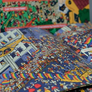 Może przedstawiać: Otwarta książka z kolorową ilustracją tłumu ludzi na paradzie. Ilustracja przedstawia ż&oacute;łty helikopter lecący nad nimi i tęczowe flagi.