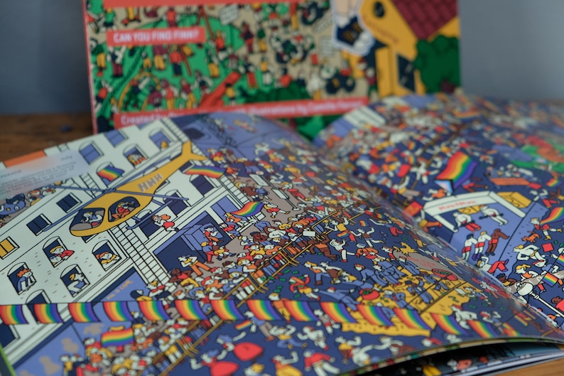 Może przedstawiać: Otwarta książka z kolorową ilustracją tłumu ludzi na paradzie. Ilustracja przedstawia ż&oacute;łty helikopter lecący nad nimi i tęczowe flagi.