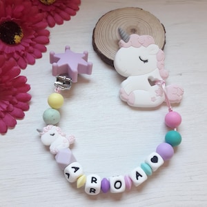 May include: A baby pacifier clip with a white and pink unicorn design, pastel beads, and the name "ARROA" spelled out in letter beads. The clip features a star-shaped clasp and is surrounded by pink flowers and a wooden slice.