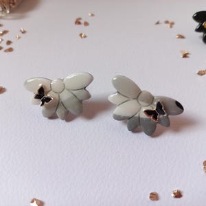 May include: A pair of gray and white ceramic earrings with black butterfly accents. The earrings are shaped like flowers with a textured surface.