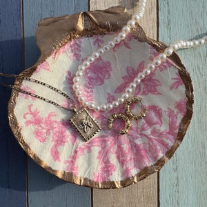 Op de afbeelding: Een witte schelpvormige schaal met een roze bloemenpatroon en gouden rand. De schaal is gevuld met een parelketting, een gouden ketting met een gouden hanger en een paar gouden oorringen.