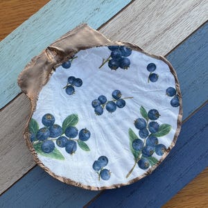 May include: A decorative seashell dish with a white interior featuring a blueberry design. The rim is painted gold, and the dish is resting on a wooden surface with blue and gray painted planks.