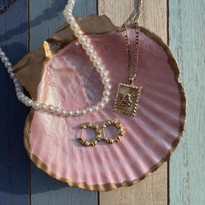 Könnte beinhalten: Eine rosa und goldene Muschelschüssel mit einer Perlenkette, einer goldenen Kettenkette mit einem Anhänger und zwei goldenen Creolen.