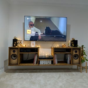 May include: A wooden entertainment unit with a floating shelf design. The shelf features a record player, speakers, and a TV mounted above it. The shelf is decorated with fairy lights and records.