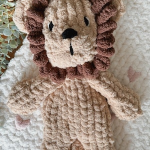 Puede incluir: Un juguete de le&oacute;n de peluche en tonos beige y marr&oacute;n. El le&oacute;n tiene un aspecto texturizado y esponjoso, con una melena marr&oacute;n y detalles faciales negros. Descansa sobre una superficie blanca y texturizada.