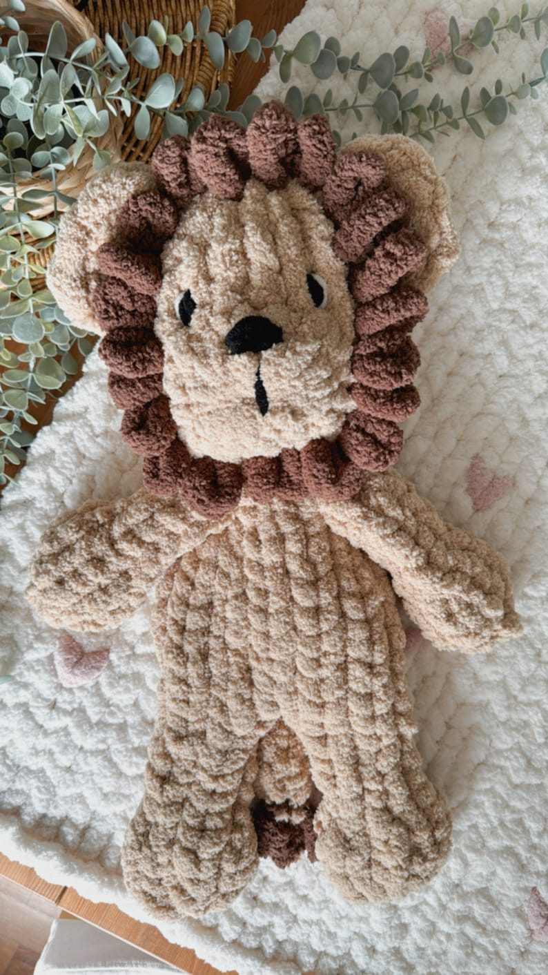 Puede incluir: Un juguete de le&oacute;n de peluche en tonos beige y marr&oacute;n. El le&oacute;n tiene un aspecto texturizado y esponjoso, con una melena marr&oacute;n y detalles faciales negros. Descansa sobre una superficie blanca y texturizada.