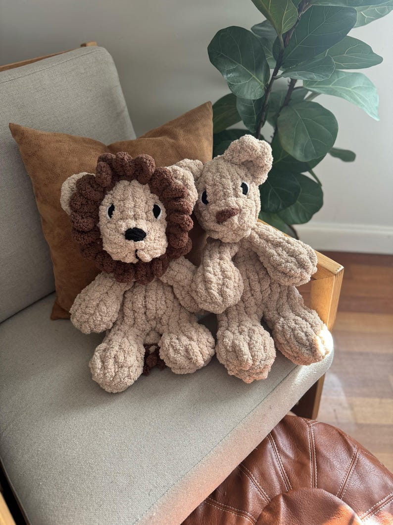 Puede incluir: Dos juguetes de peluche de le&oacute;n hechos a mano. Un le&oacute;n tiene una melena marr&oacute;n y el otro una cabeza simple. Ambos son beige y tienen ojos y narices negros. Est&aacute;n sentados en una silla gris claro con un coj&iacute;n marr&oacute;n.