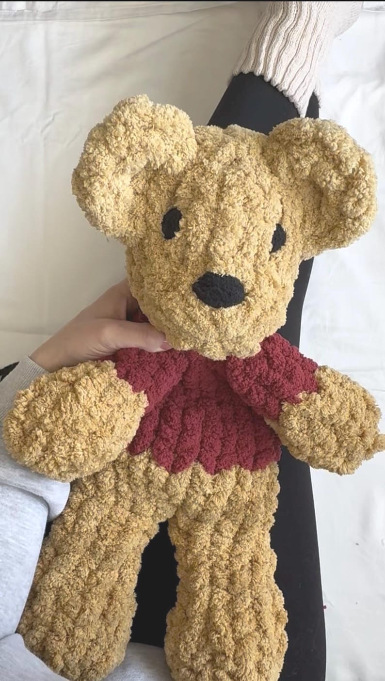 Puede incluir: Un oso de peluche hecho a mano, con un cuerpo y una cabeza de color marr&oacute;n claro texturizado. El oso tiene un su&eacute;ter rojo oscuro y ojos y nariz negros. El juguete blando est&aacute; dise&ntilde;ado con orejas y extremidades grandes y redondeadas.