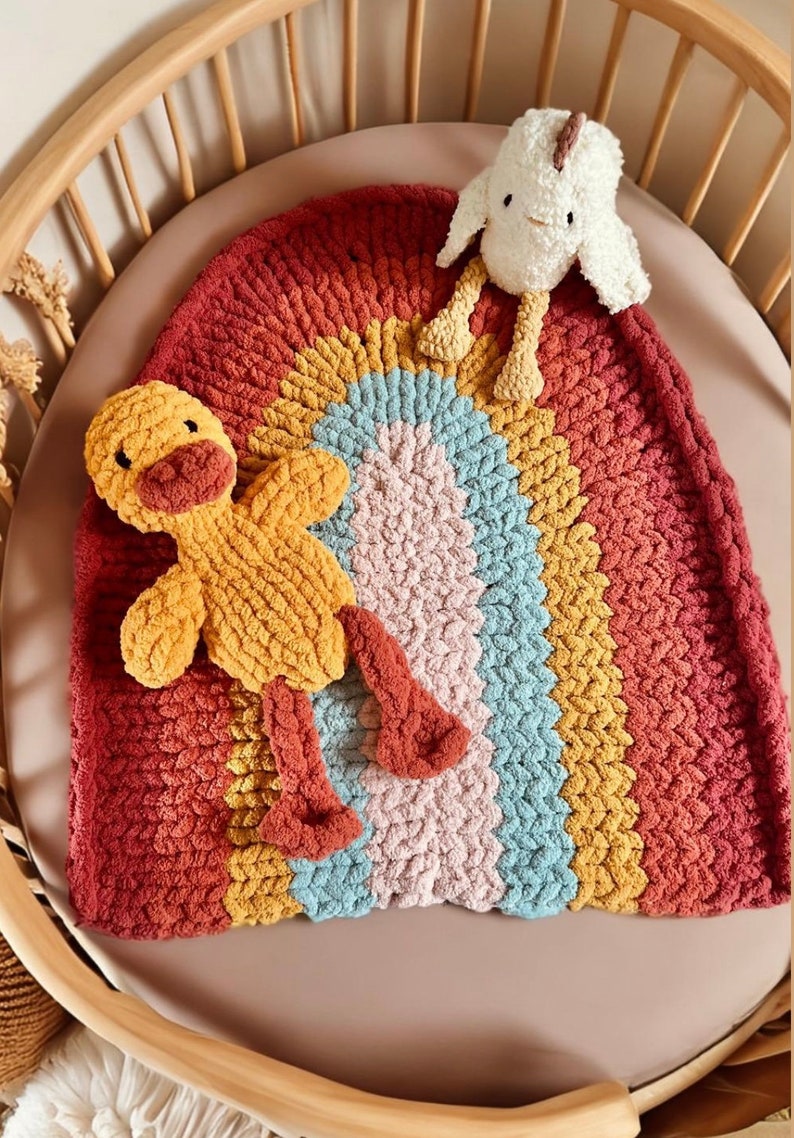 May include: A rainbow-colored crocheted baby blanket with a yellow duck and a white chick plush toy. The blanket is made of chunky yarn and has a soft, textured feel.
