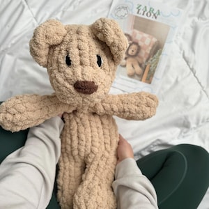 Puede incluir: Un oso de peluche tejido a mano en color beige claro, con una nariz marr&oacute;n oscuro y ojos negros. El oso tiene los brazos extendidos y est&aacute; hecho de un hilo suave y texturizado. Un libro de patrones titulado "ZARA LION" es visible en el fondo.