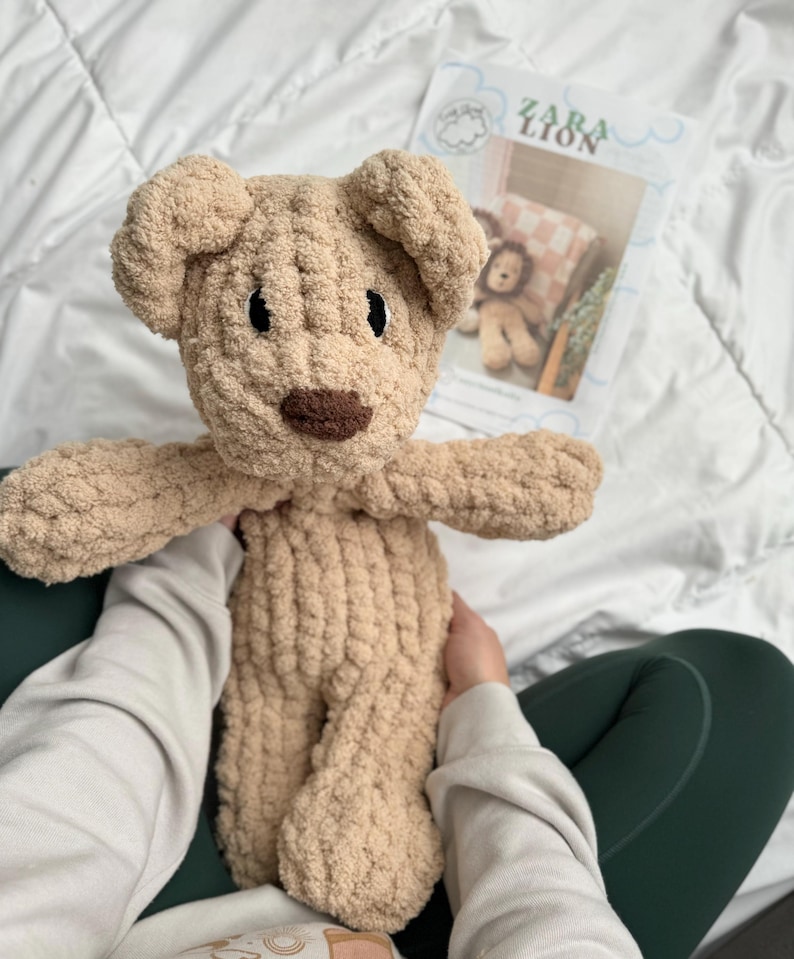 Puede incluir: Un oso de peluche tejido a mano en color beige claro, con una nariz marr&oacute;n oscuro y ojos negros. El oso tiene los brazos extendidos y est&aacute; hecho de un hilo suave y texturizado. Un libro de patrones titulado "ZARA LION" es visible en el fondo.