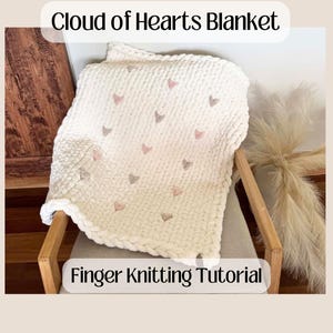 Può includere: Una coperta a maglia grossa color crema con cuori rosa e grigi ricamati, drappeggiata su una sedia di legno. La coperta è etichettata "Cloud of Hearts Blanket" e "Finger Knitting Tutorial".