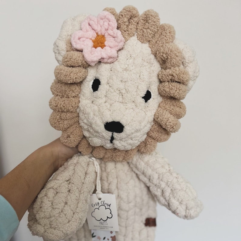 Puede incluir: Un juguete de le&oacute;n de peluche hecho a mano, con un cuerpo color crema y una melena marr&oacute;n claro. El le&oacute;n tiene ojos negros y una nariz negra. Una flor rosa con un centro naranja adorna la parte superior de su cabeza. Una etiqueta est&aacute; adjunta al juguete.