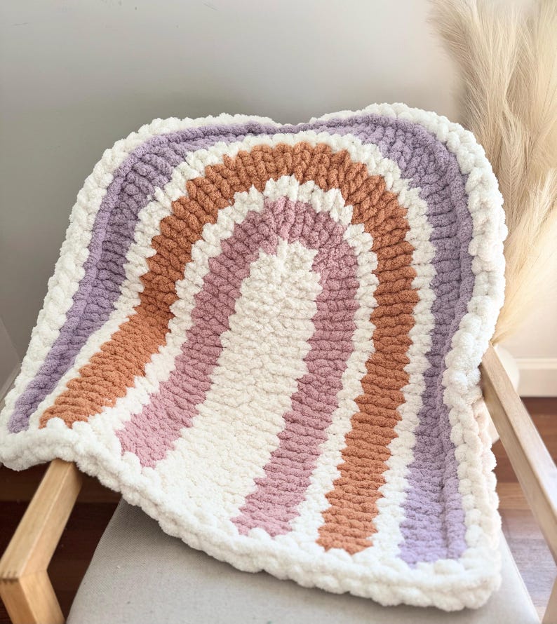 May include: A crocheted rainbow blanket with stripes of purple, orange, pink, and white. The blanket is made with a fluffy yarn and has a soft, textured look.