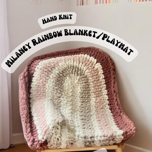 May include: A hand-knitted rainbow blanket or playmat in shades of pink, white, and brown. The blanket is draped over a wooden chair with the text "MILANEY RAINBOW BLANKET/PLAYMAT" and "HAND KNIT" above it. The text "VIDEO TUTORIAL" is below the blanket.