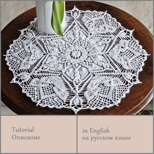 May include: A white, intricately crocheted doily with a floral and geometric design. The doily is circular, with a scalloped edge and a central floral motif. The doily is placed on a dark wooden table, with a white vase and green leaf in the background.
