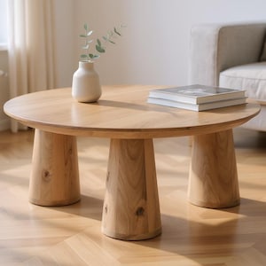 Könnte beinhalten: Ein runder Couchtisch aus hellem Holz mit drei konischen Beinen. Auf dem Tisch stehen eine Vase mit Grün und zwei Bücher. Der Tisch steht auf einem Fischgrät-Parkett.