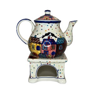 Può includere: Una teiera in ceramica con scaldavivande abbinato. La teiera è bianca con pois colorati e una scena di villaggio. Lo scaldavivande ha un design abbinato con una piccola apertura. La teiera ha un manico e un beccuccio blu.