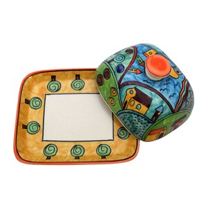 May include: A ceramic butter dish with a colorful, hand-painted design of a whimsical town with houses, trees, and a blue sky. The dish has a square base and a domed lid with a small orange knob.
