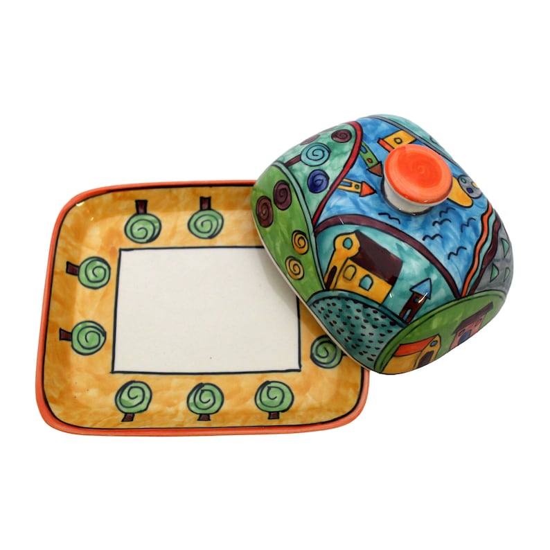 May include: A ceramic butter dish with a colorful, hand-painted design of a whimsical town with houses, trees, and a blue sky. The dish has a square base and a domed lid with a small orange knob.