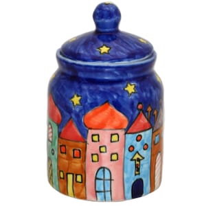 Op de afbeelding: Een blauwe keramische pot met een deksel, versierd met een nachtelijke hemel met sterren en kleurrijke huizen. De huizen hebben rode, oranje, roze en blauwe accenten.