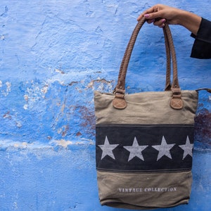 Può includere: Una borsa tote in tela beige con stelle bianche e nere e il testo "Vintage Collection". La borsa ha manici in pelle marrone.