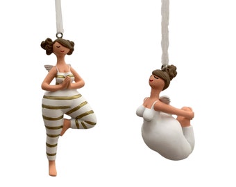 May include: Two white ceramic ornaments of women in yoga poses. One woman is in a tree pose with gold stripes on her white pants. The other woman is in a seated forward bend pose. Both women have brown hair and are wearing white tops.