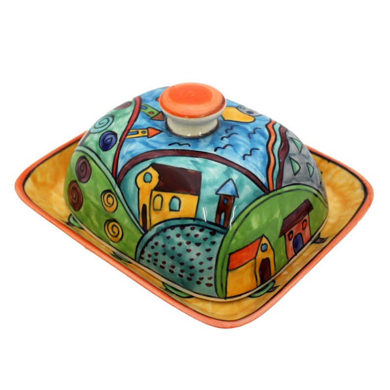 May include: A ceramic butter dish with a colorful, whimsical design depicting a village with houses, trees, and a blue sky. The dish has a lid with a bright orange knob.