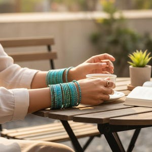 Könnte beinhalten: Eine Person trägt mehrere türkisfarbene und blaue Armreifen an jedem Handgelenk und hält eine weiße Teetasse. Die Armreifen bestehen aus Glas oder einem ähnlichen Material. Die Person sitzt draußen an einem Holztisch.