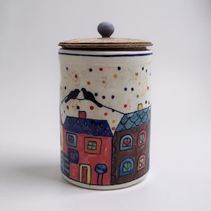 Puede incluir: Un tarro de cerámica con tapa, con un diseño pintado a mano de casas y pájaros. El tarro es cilíndrico con una base blanca, una tapa marrón y un pequeño pomo azul. Las casas son rojas y marrones con tejados azules. El fondo está salpicado de puntos.
