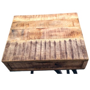 May include: A square, wooden side table with a natural finish. The tabletop is made of planks and has a drawer. The table is supported by black metal legs. The table measures approximately 51 cm wide and 51 cm deep.