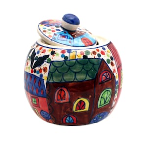 Könnte beinhalten: Ein handbemalter Keramik-Zuckertopf mit Deckel, der eine skurrile Dorfszene zeigt. Die Schale zeigt braune, rote und blaue Häuser mit grünen Dächern und bunten Fenstern. Der Deckel hat einen blauen Knopf und ähnliche Motive.