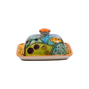 May include: A colorful ceramic butter dish with a lid. The dish features a whimsical design of green hills, blue sky, and orange houses.