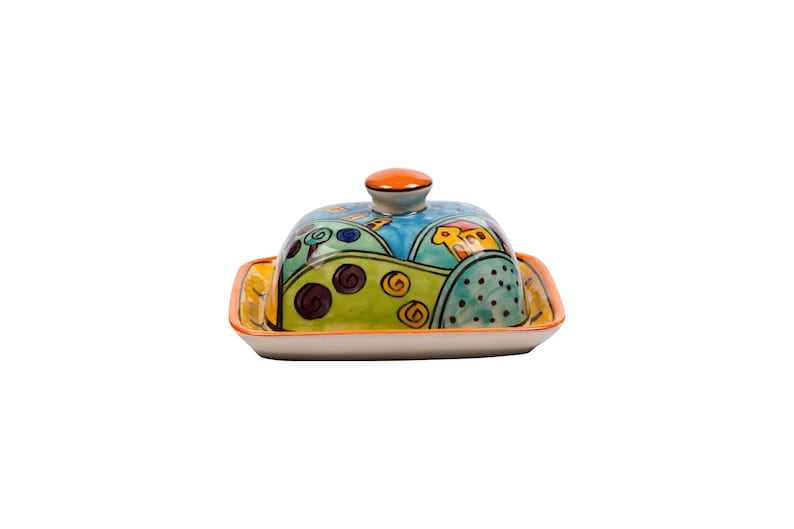 May include: A colorful ceramic butter dish with a lid. The dish features a whimsical design of green hills, blue sky, and orange houses.
